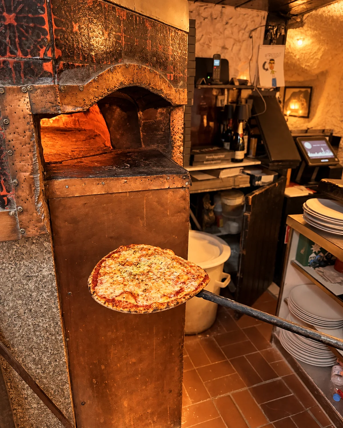 Eine dünne Pizza kommt auf dem Schieber aus dem Holzofen